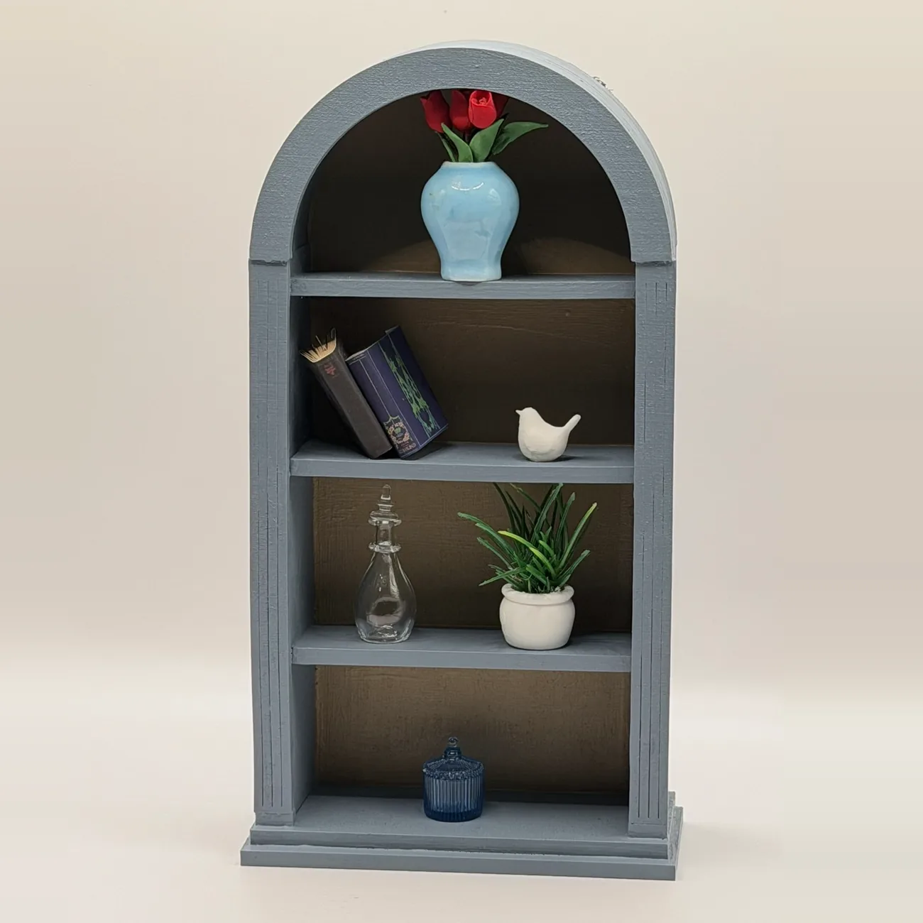 The Wren Arched Bookcase, a blue-gray miniature bookcase with an arched top and four open shelves, styled with tiny books, a vase of tulips, and a potted plant.